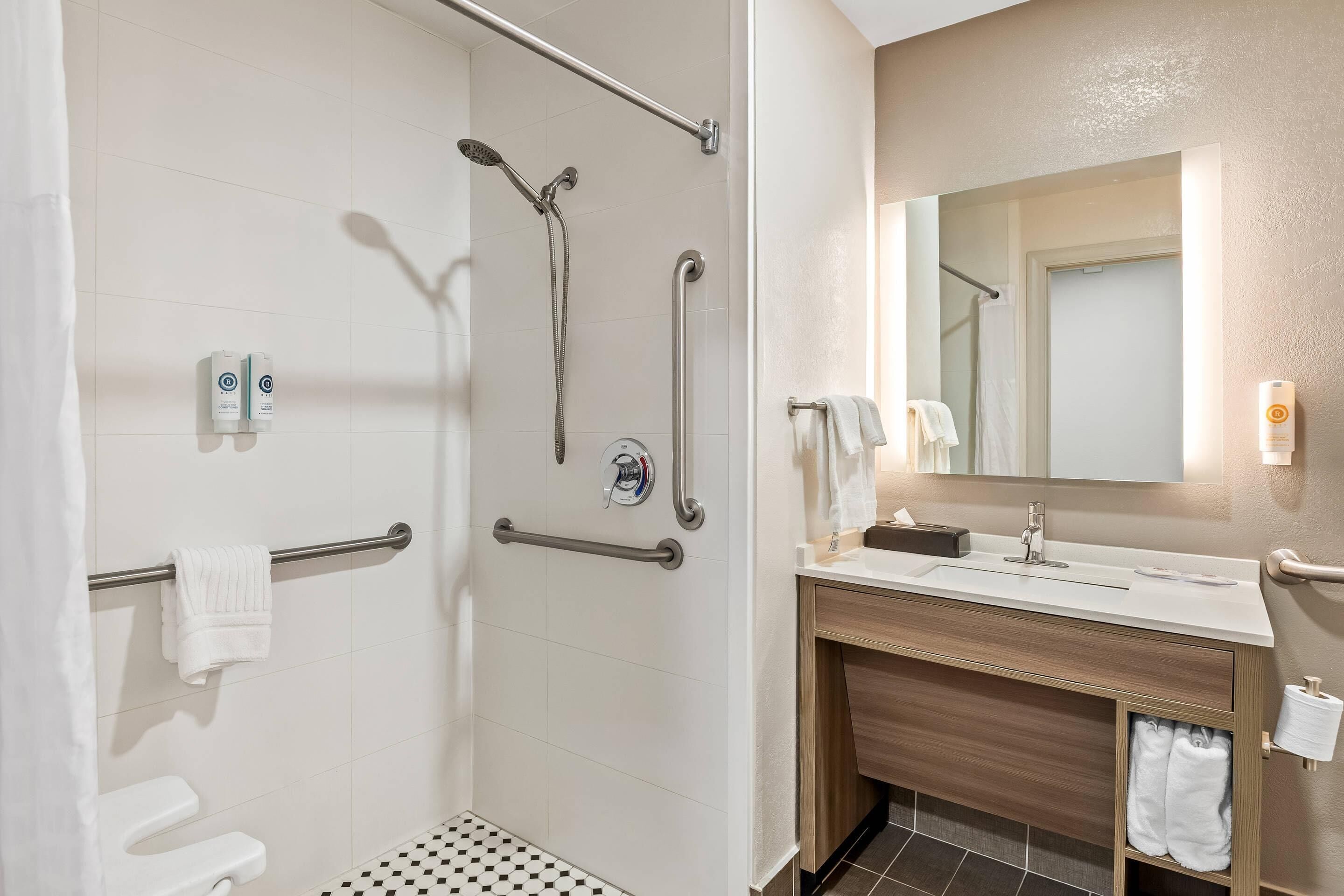 A modern bathroom with a shower, grab bars, a toilet, and a sink with a mirror.