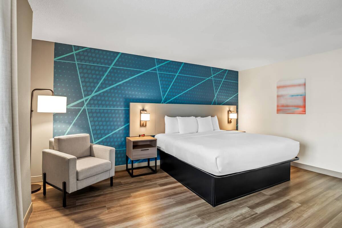 A modern hotel room with a large bed, a chair, a nightstand, and a wall with a geometric pattern.