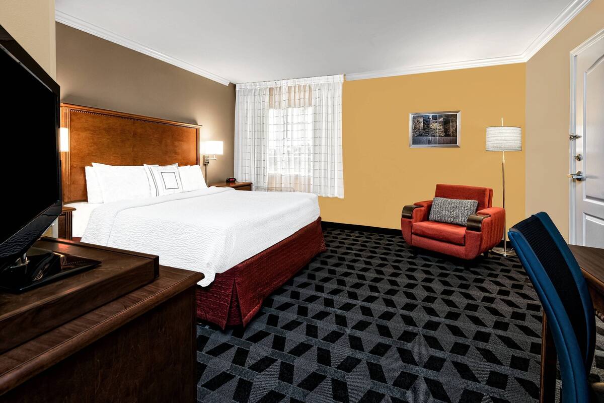 A hotel room with a large bed, a desk with a telephone, a red armchair, a floor lamp, and a painting on the wall.