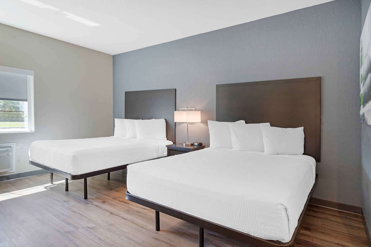 A hotel room with two single beds, a bedside table with a lamp, and a large window providing a view of the outdoors.