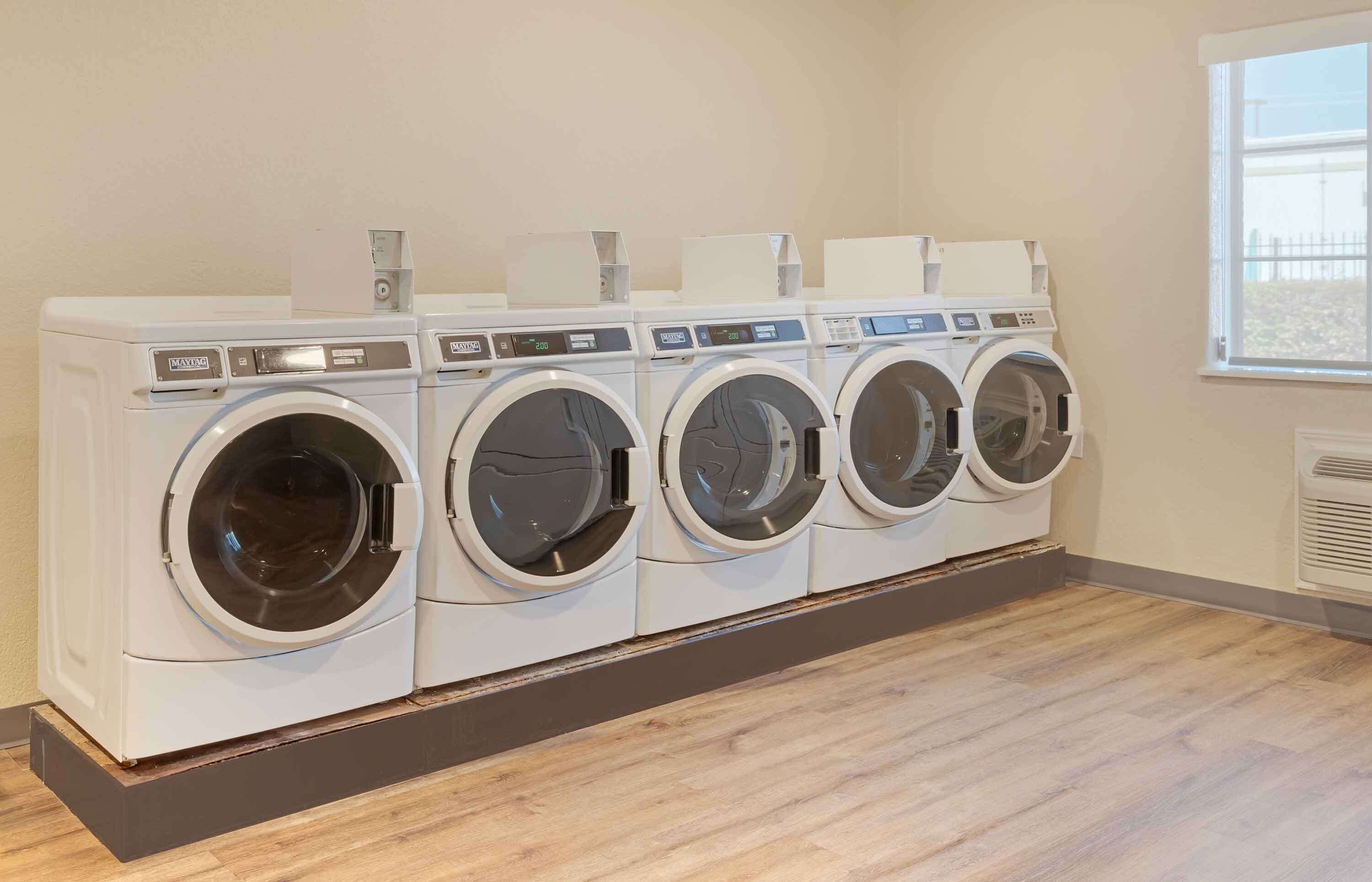 A row of four front-loading washing machines in a laundry room with a wooden floor and a window in the background.