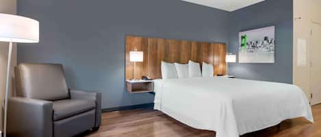 A hotel room with a bed, a chair, a nightstand, and a wall-mounted shelf.