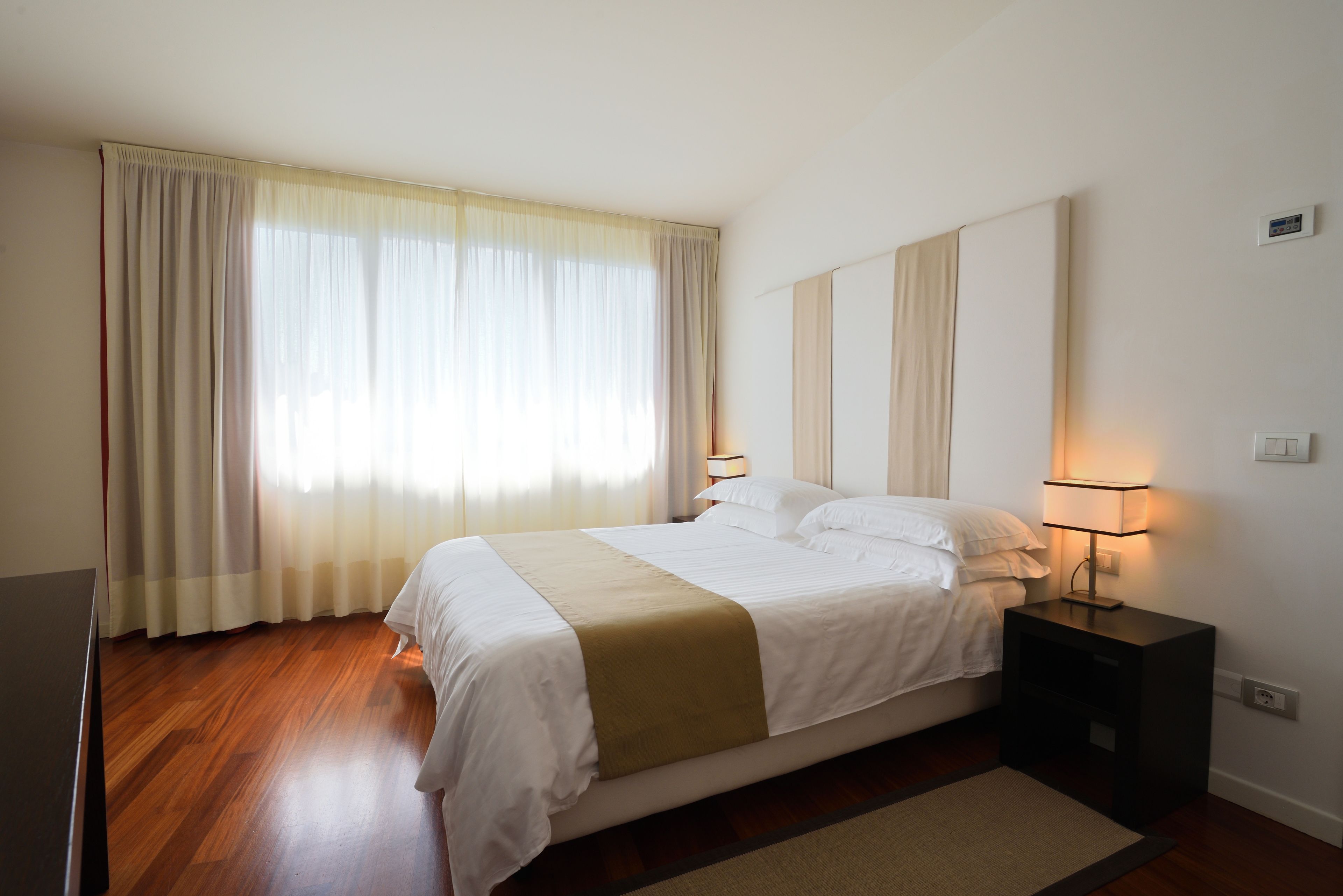 A neatly made bed with a white and beige bedspread, a bedside table with a lamp, and a large window with sheer curtains.
