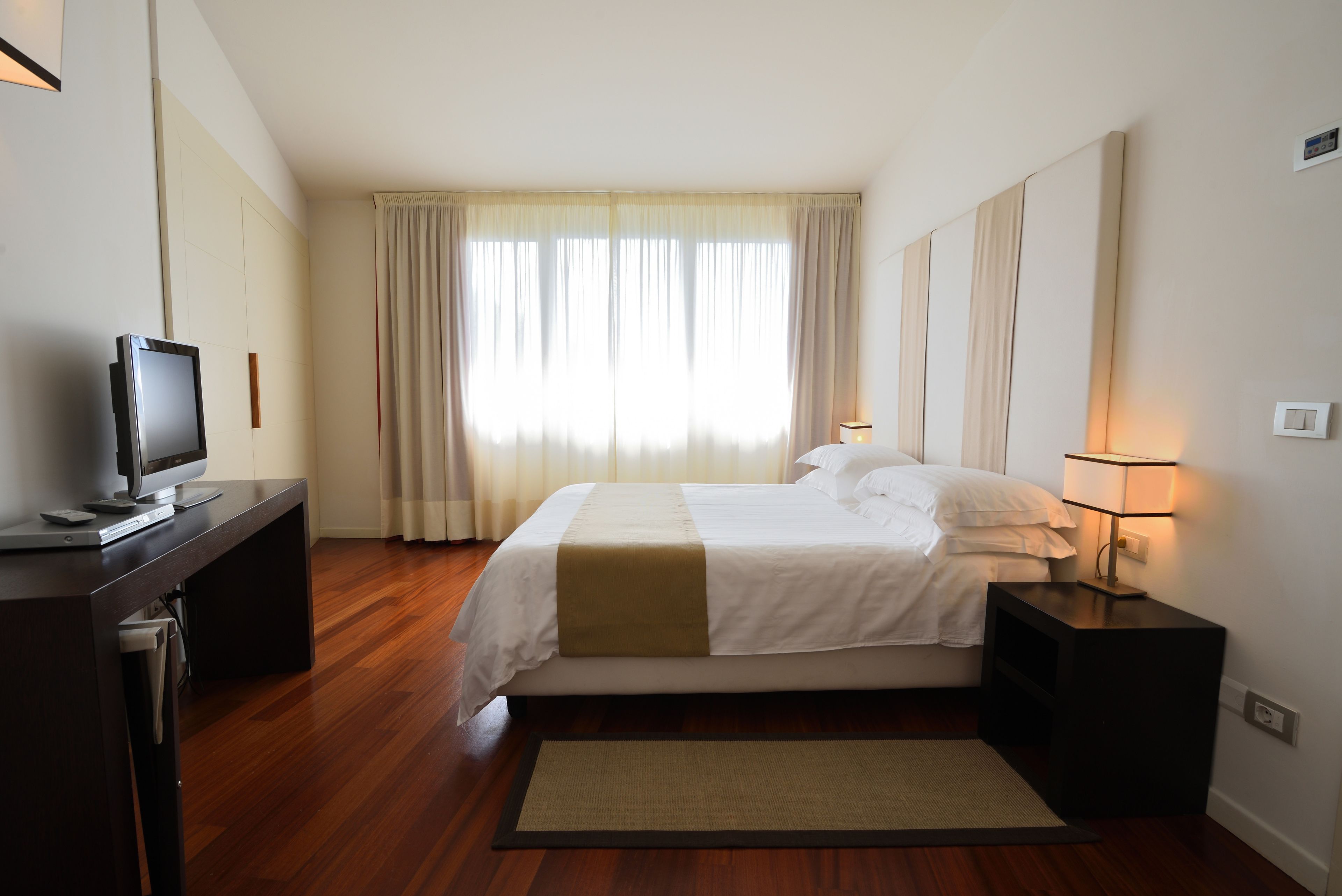 A hotel room with a bed, a desk with a computer, a lamp, and a window with curtains.
