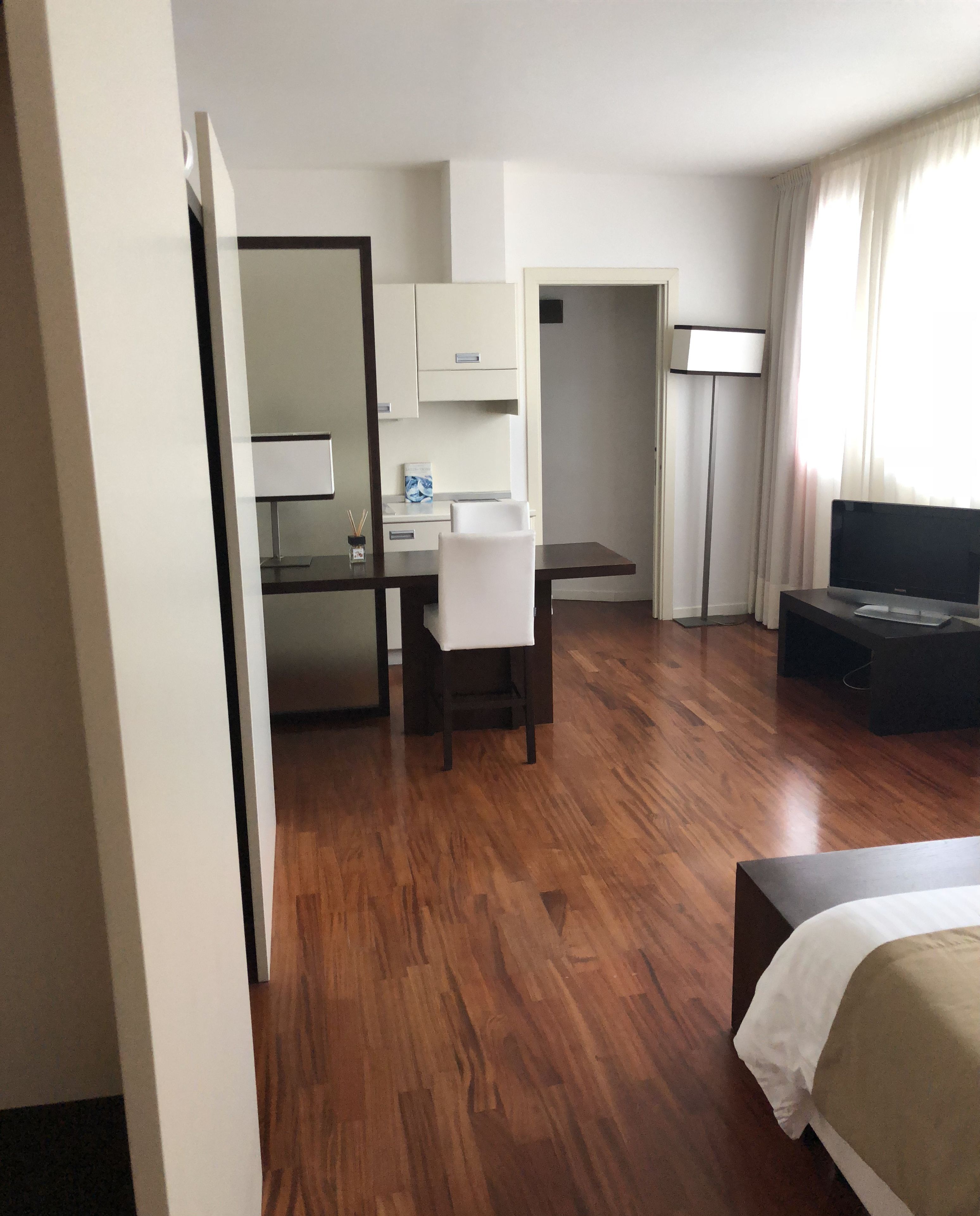 Une chambre d’hôtel avec un plancher en bois, un lit, un bureau avec une chaise, une télévision et une fenêtre avec des rideaux.