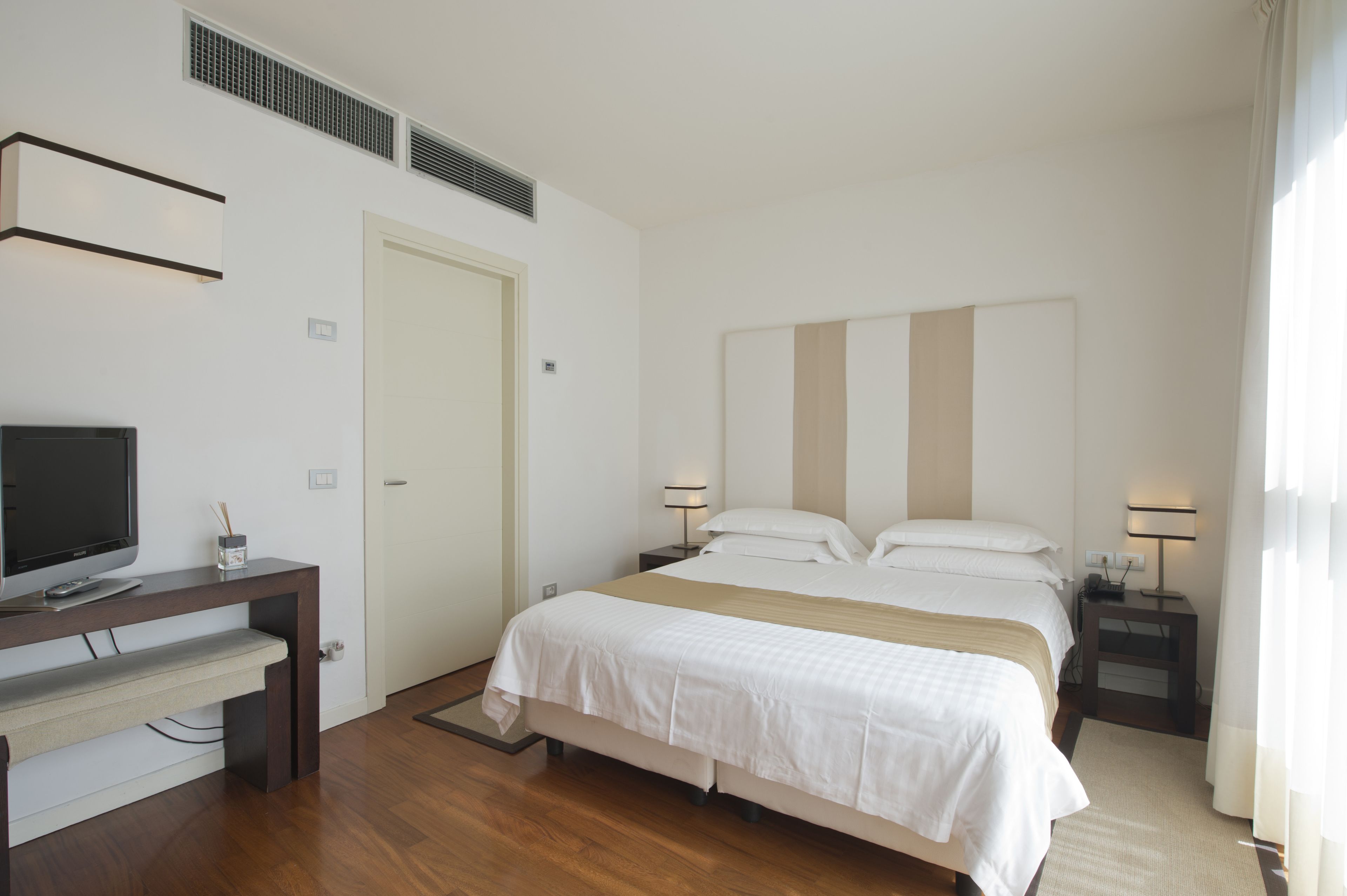 A hotel room with a bed, bedside tables, a desk, a television, and a window with curtains.