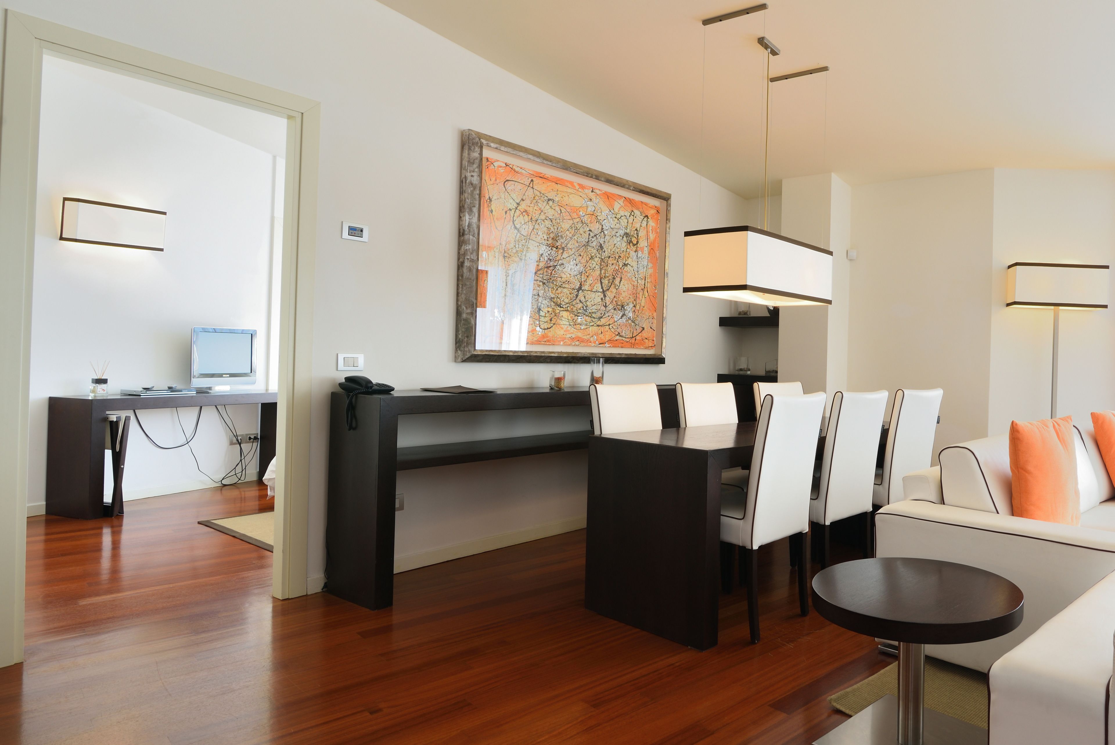 A modern living room with a dining area, a large abstract painting on the wall, and a flat-screen TV on a desk.