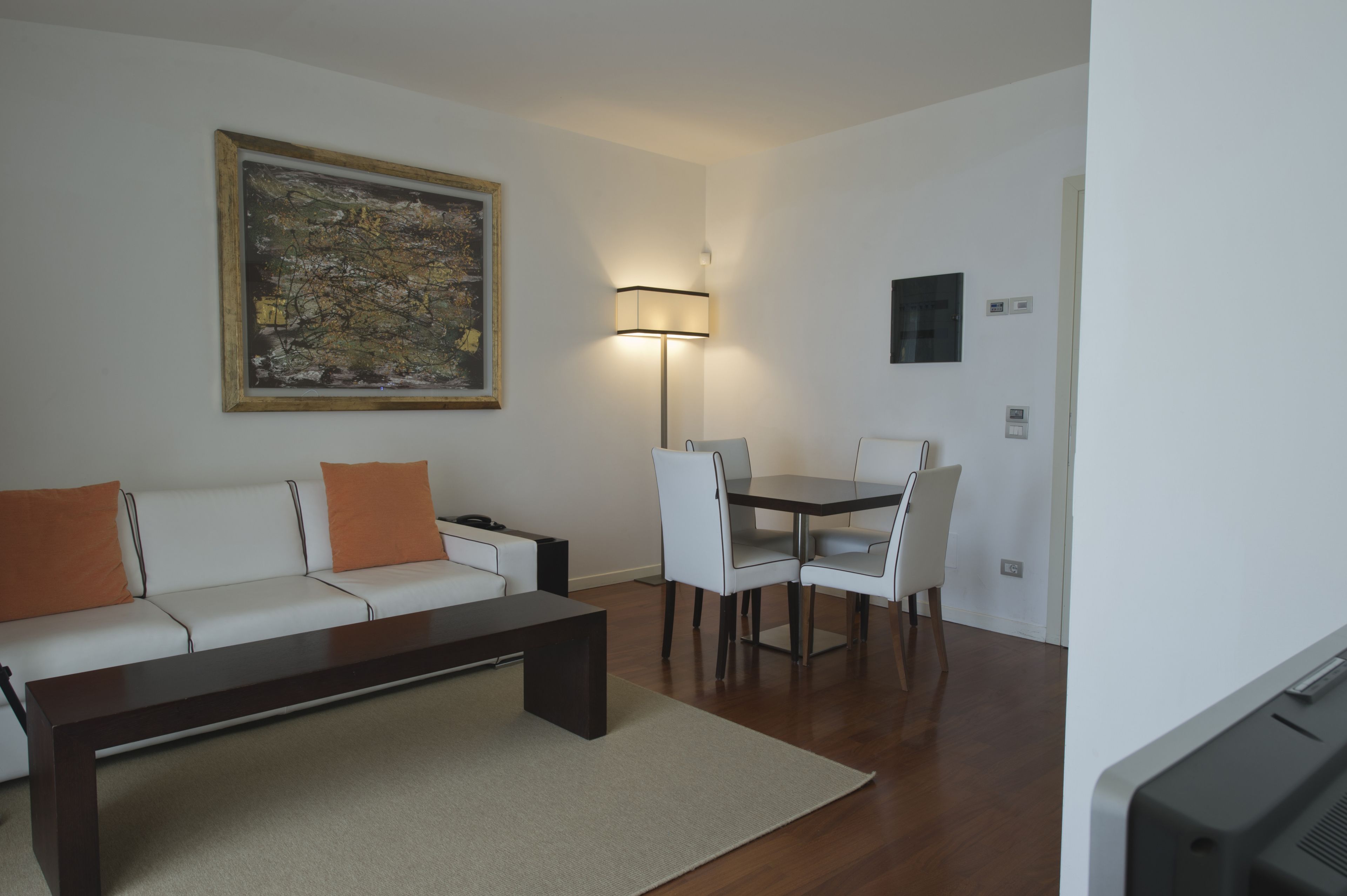 Ein modernes Wohnzimmer mit einem weißen Sofa, einem dunklen Couchtisch und einem Essbereich inklusive Tisch und Stühlen. Ein großes abstraktes Gemälde ist an der Wand aufgehängt, und eine Stehlampe befindet sich in der Nähe des Essbereichs.