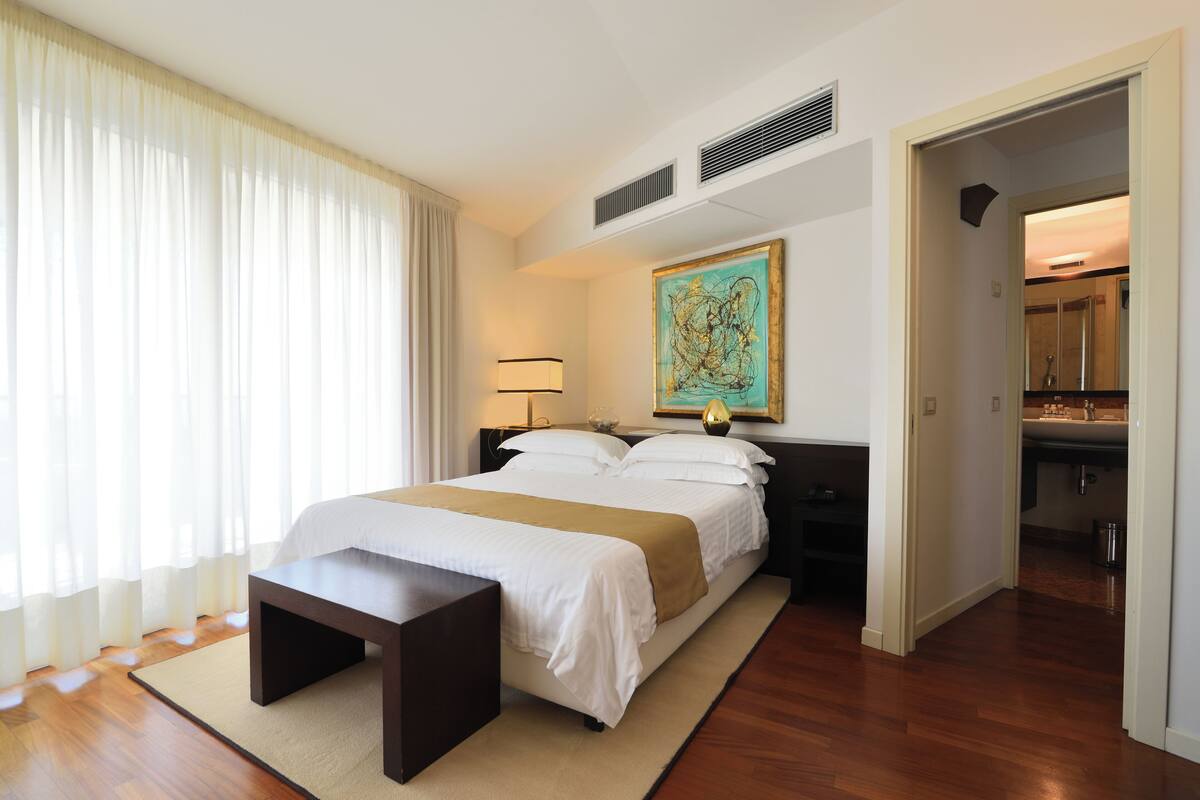 Habitación de hotel con una cama grande, mesitas de noche, un banco, un cuadro y vista al baño.