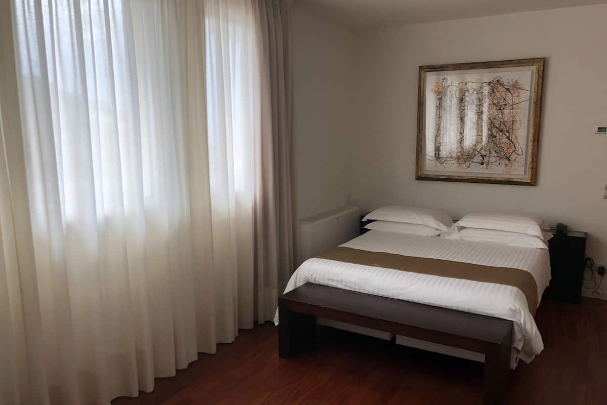Un dormitorio con cama, un banco, un cuadro en la pared y una ventana con cortinas.