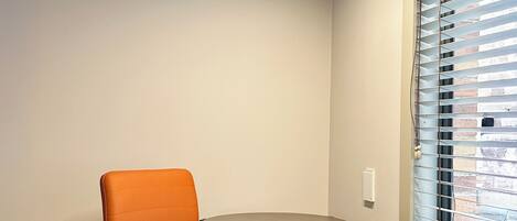Un espacio de oficina moderno con una mesa redonda, una silla de oficina color naranja, una ventana con persianas y un cesto de basura.