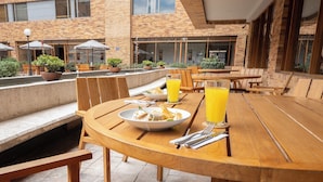 Un área de comedor exterior con mesas y sillas de madera, un edificio de ladrillo al fondo y un cielo despejado.