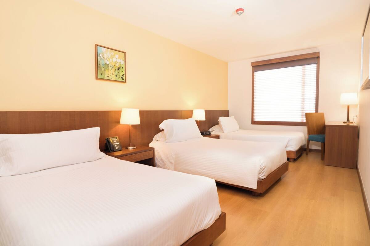 A hotel room with three beds, wooden headboards, bedside lamps, a small desk, and a window with blinds.