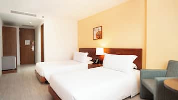 A hotel room with two beds, a small table, a chair, and a wall-mounted lamp.
