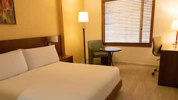 A hotel room with a large bed, a desk, a chair, a lamp, and a window with blinds.