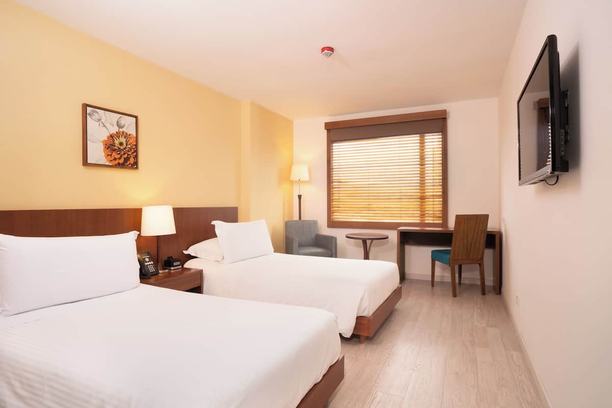 A hotel room with two beds, a desk, a chair, a lamp, and a TV mounted on the wall.