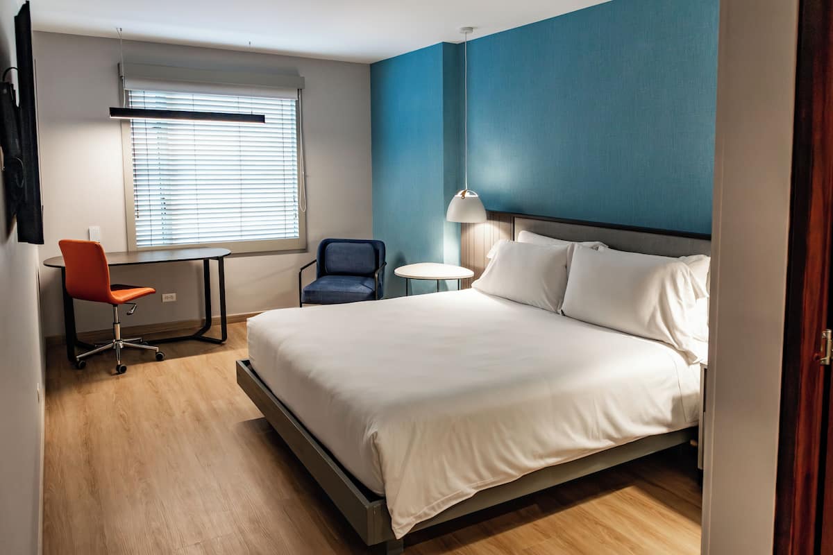 A hotel room with a large bed, a desk with a chair, a lamp, and a window with blinds.