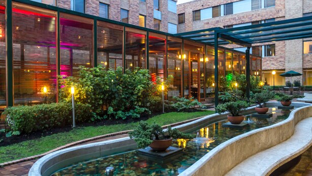 Un edificio moderno con fachada de vidrio, un jardín ajardinado con fuente de agua y zonas de estar al aire libre.