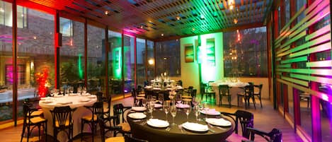 A restaurant with round tables and chairs, large windows, and a ceiling with colorful lighting.
