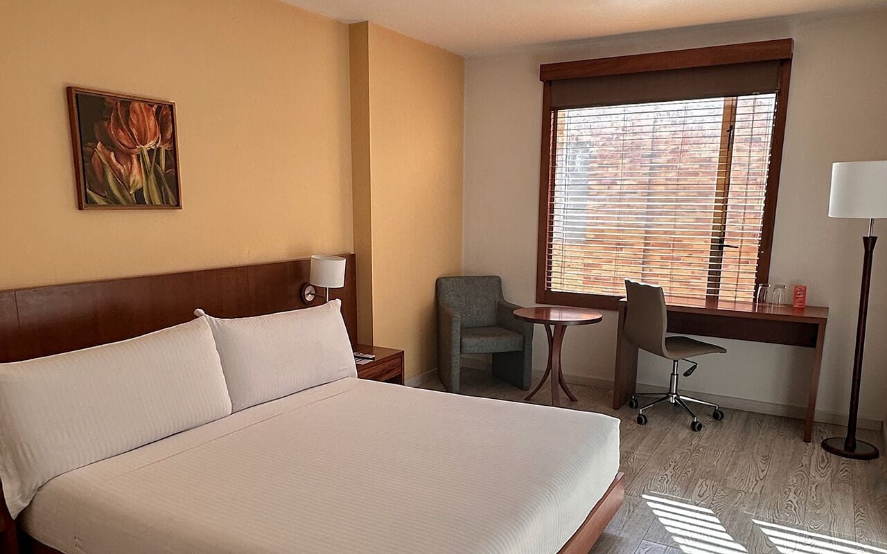 Habitación de hotel con una cama grande, un escritorio, una silla, una mesita, una lámpara y una ventana con persianas.