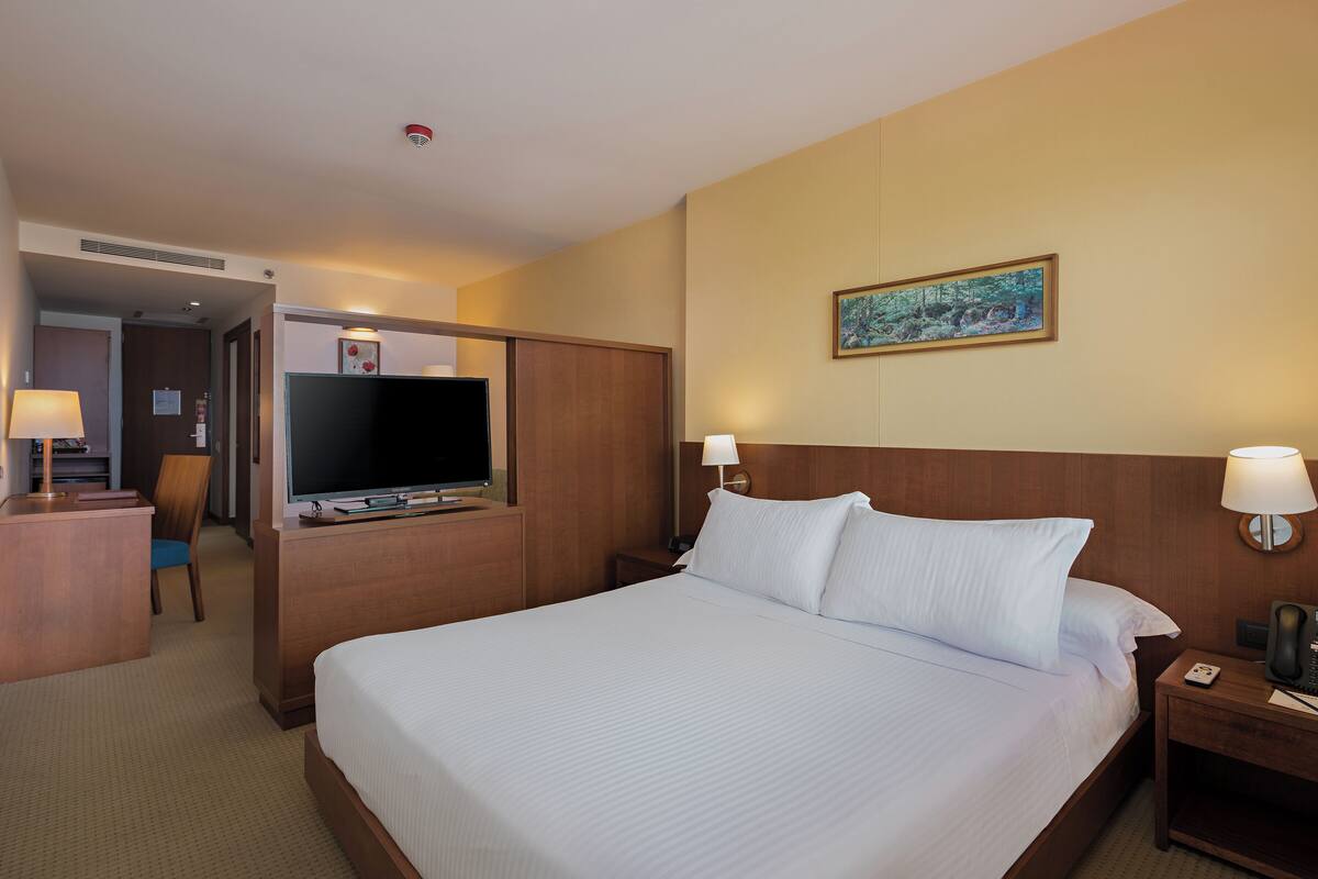 A hotel room with a large bed, a desk with a TV, a chair, and a painting on the wall.
