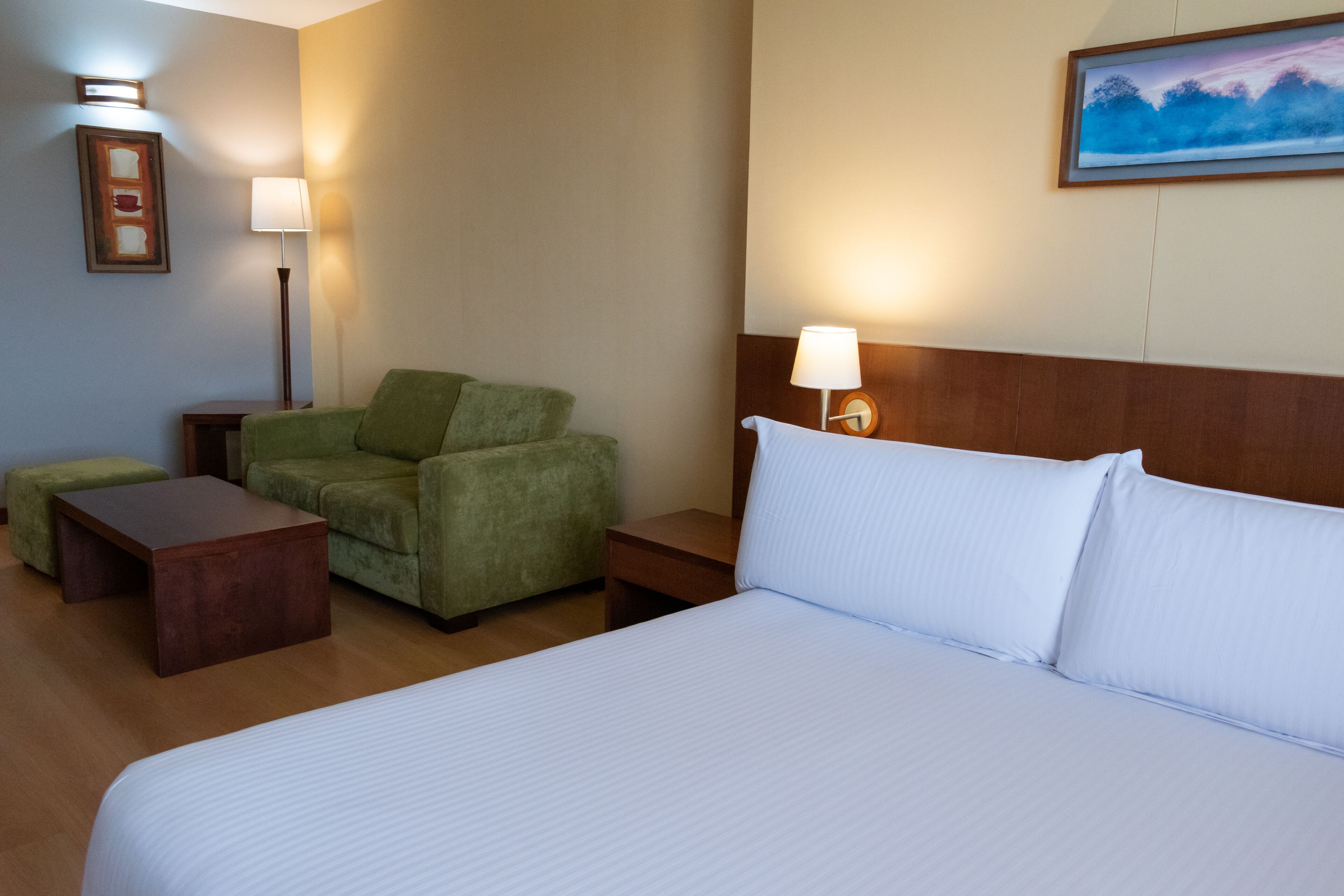 Une chambre d’hôtel avec un lit, un fauteuil vert, une table de chevet en bois, une petite table et une lampe fixée au mur.