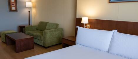 A hotel room with a bed, a green armchair, a wooden nightstand, a small table, and a wall-mounted lamp.