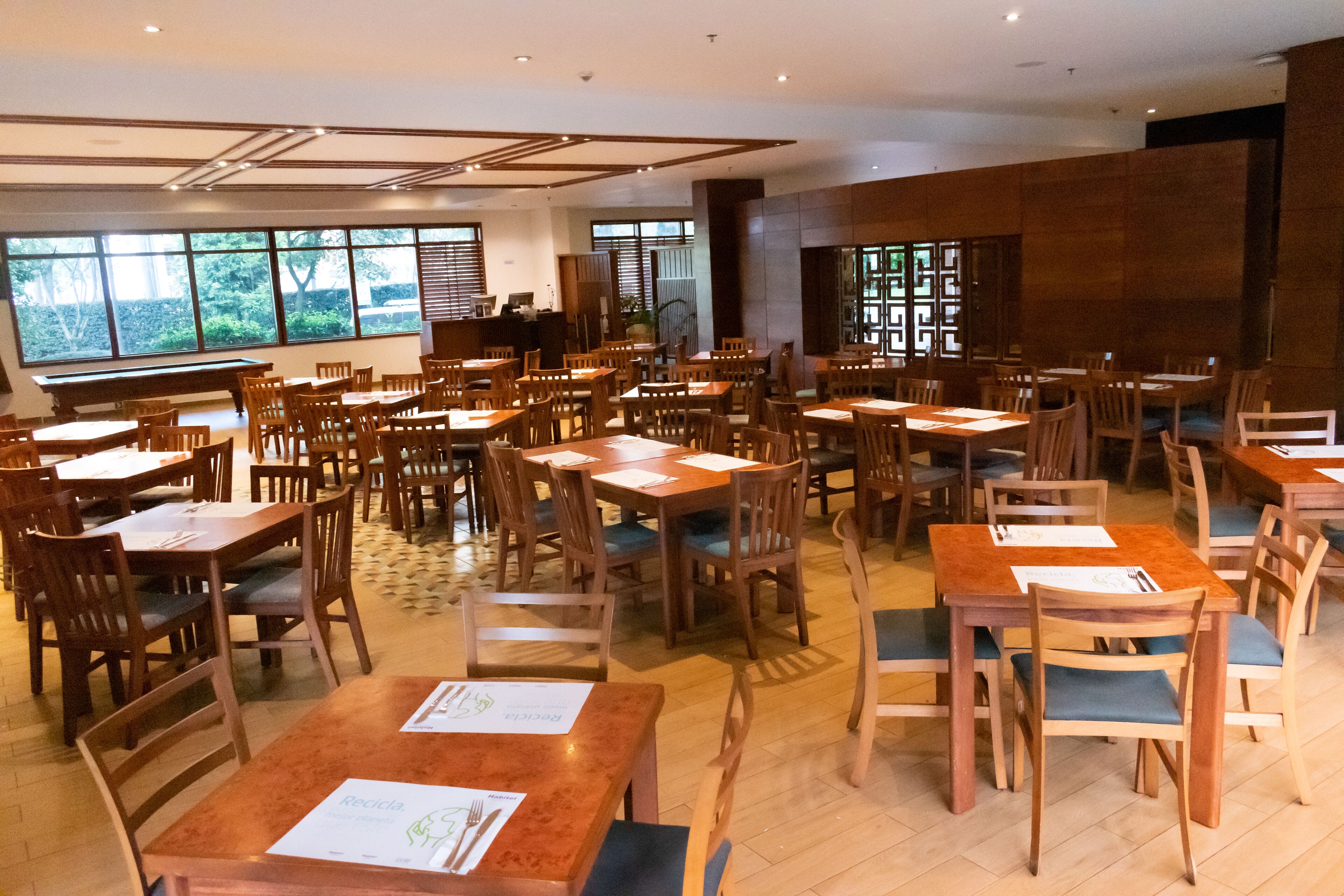 A spacious restaurant with multiple wooden tables and chairs, large windows with a view of greenery, and a modern interior design.