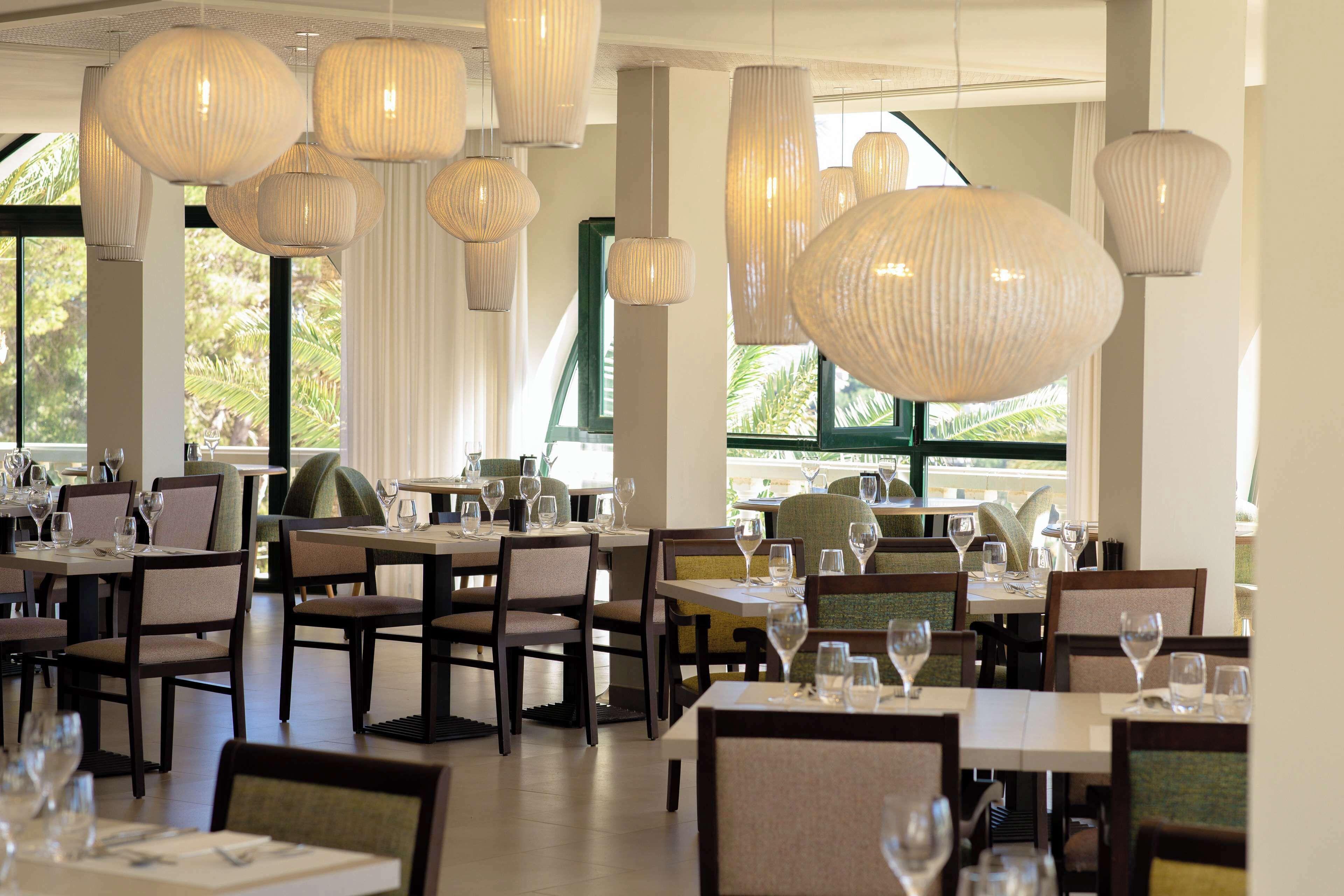 Un restaurante con varias mesas y sillas, grandes lámparas colgantes y amplias ventanas con cortinas.