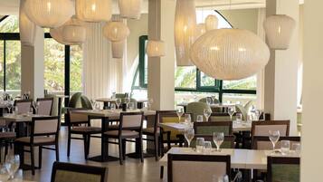 Un restaurante con varias mesas y sillas, grandes lámparas colgantes y amplias ventanas con cortinas.