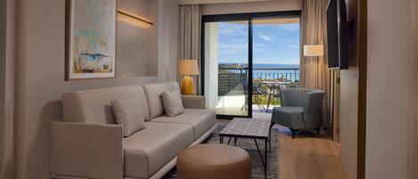 A modern hotel room with a sofa, ottoman, and a small table. There is a large window offering a view of the sea and buildings.