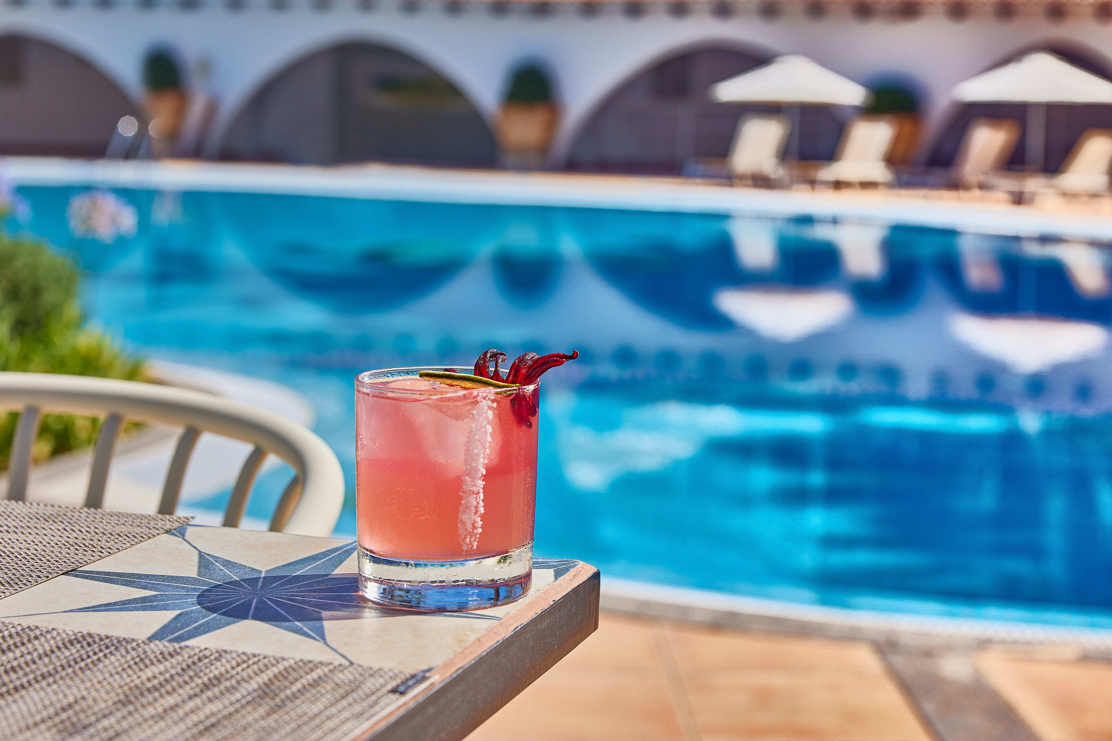 Un verre d’une boisson rose sur une table près d’une piscine, avec des chaises longues et des parasols en arrière-plan.