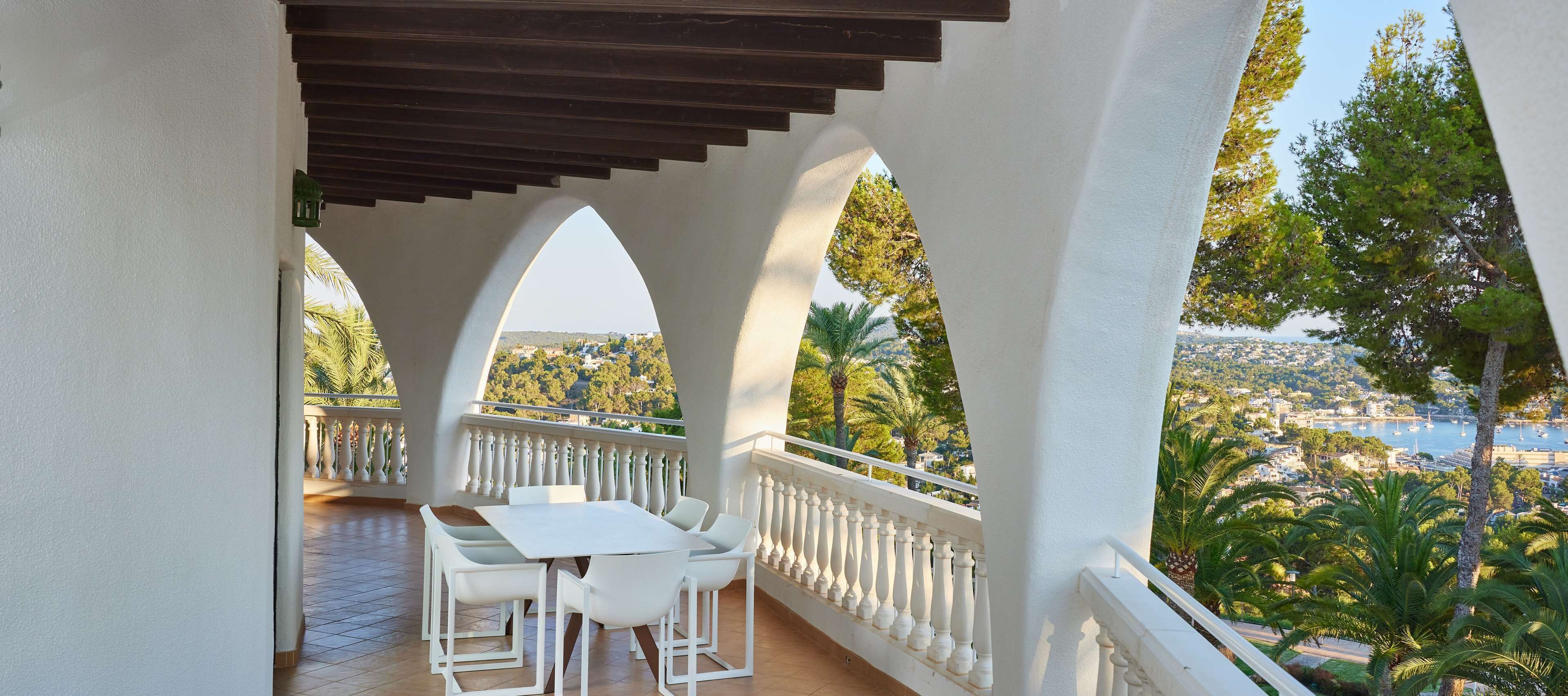 A balcony with a white table and chairs, a white balustrade, and a view of a coastal town.