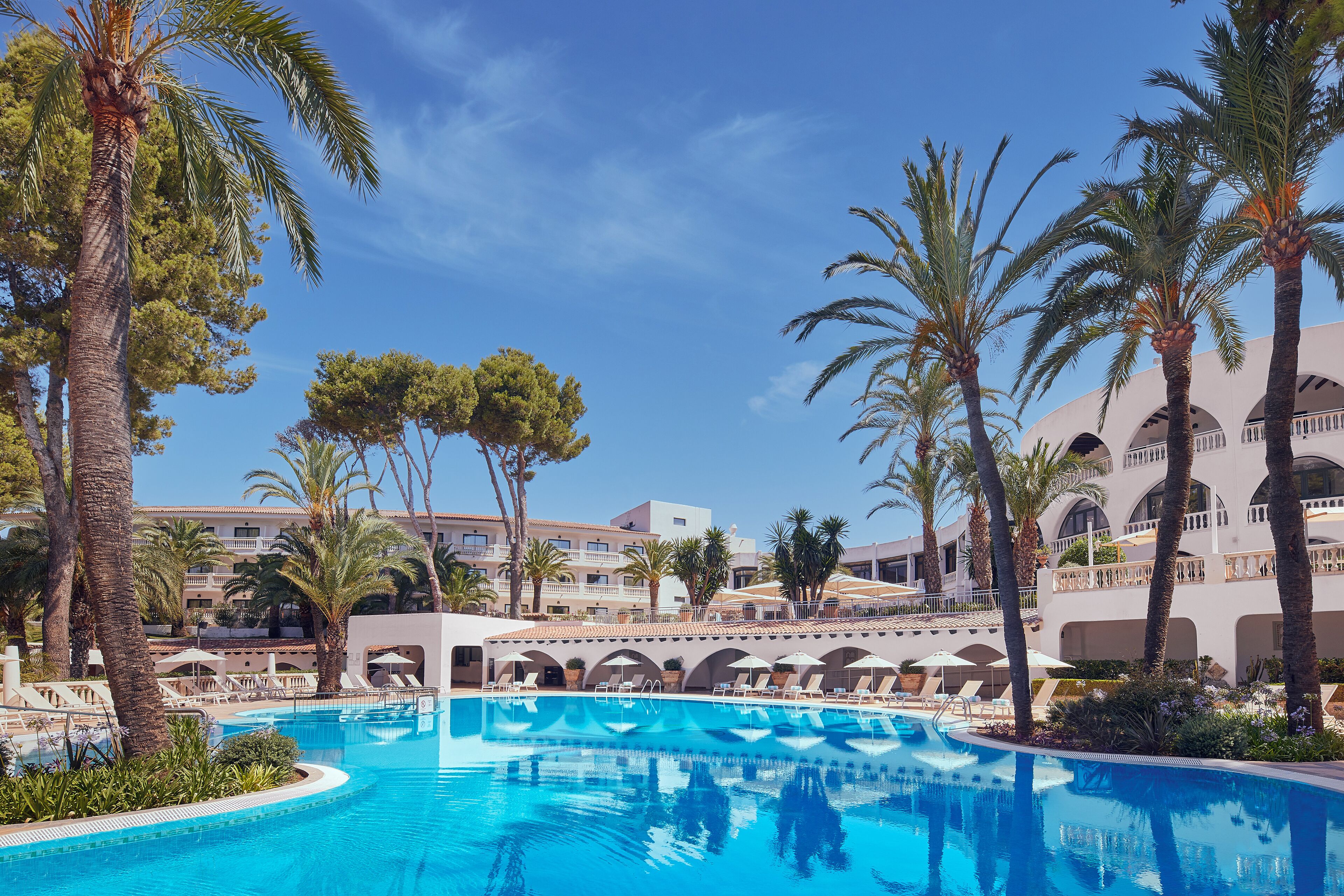 A resort pool area with palm trees, lounge chairs, and a building with arches in the background.