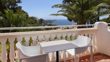 Un balcón con una mesa blanca y dos sillas, con vistas a la costa, palmeras y el mar.