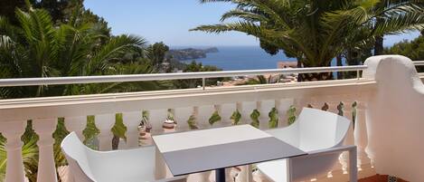 A balcony with a white table and two chairs, overlooking a coastal view with palm trees and the sea.