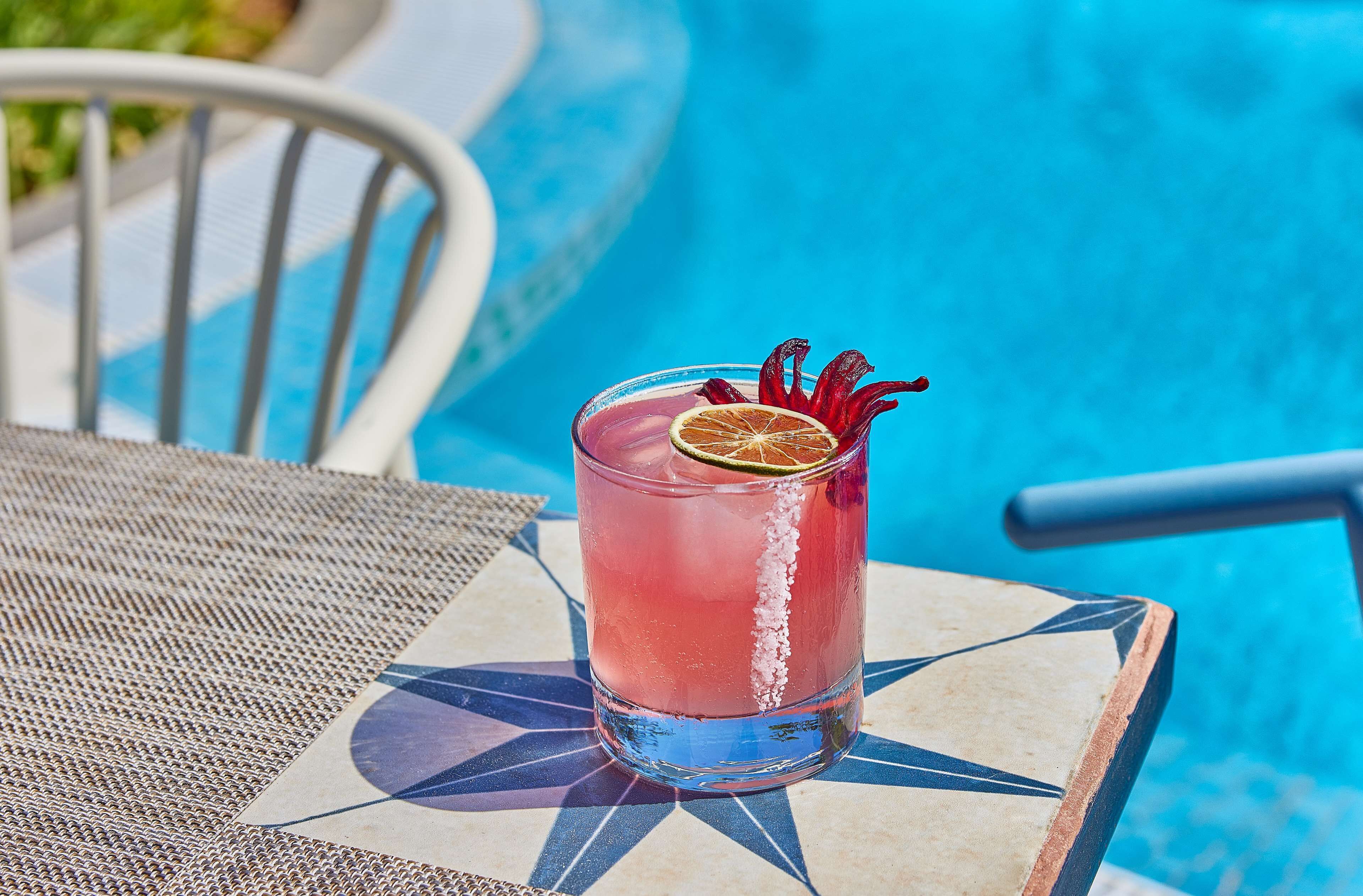 Un vaso de bebida rosa con una rodaja de lima y una guarnición roja sobre una mesa junto a una piscina.