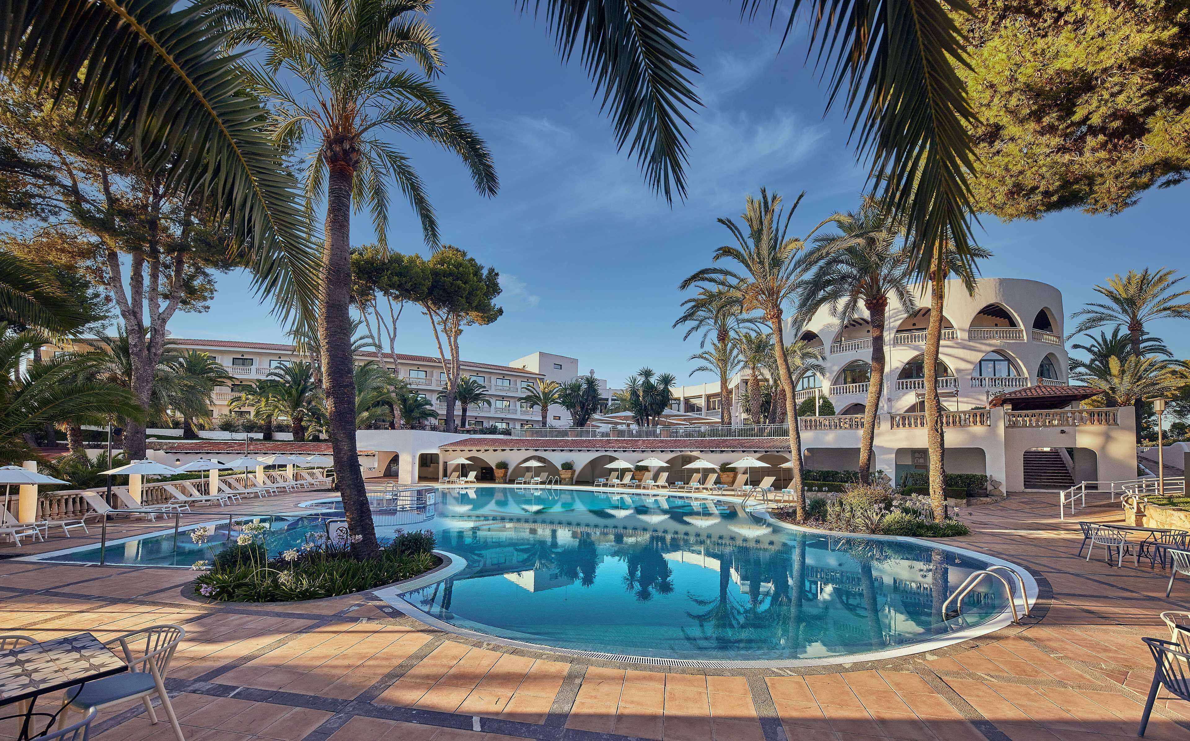 A pool area with lounge chairs and umbrellas, surrounded by palm trees and a building with arched balconies.
