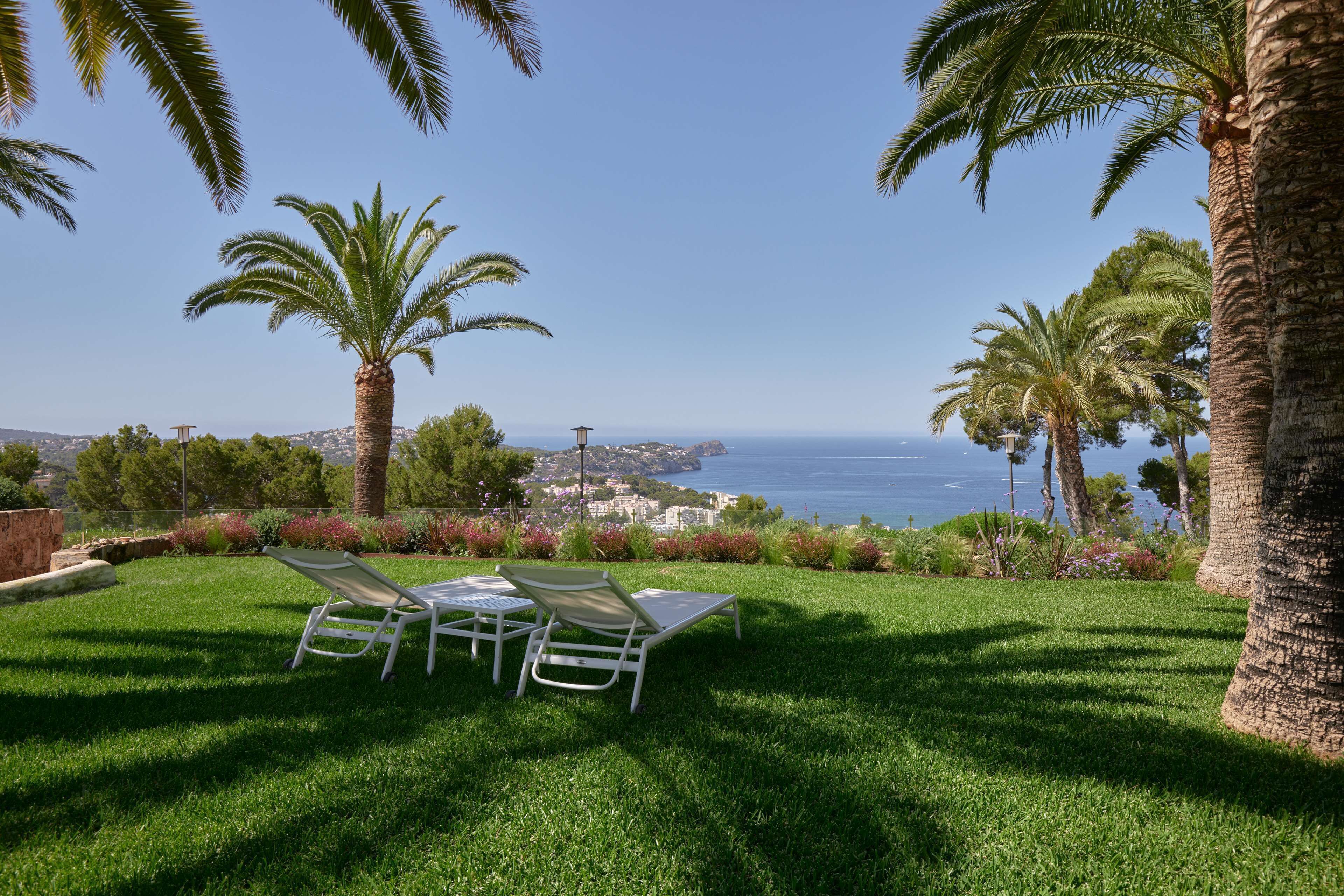 Zona verde con dos sillones blancos orientados hacia una vista panorámica del océano y un pueblo lejano.