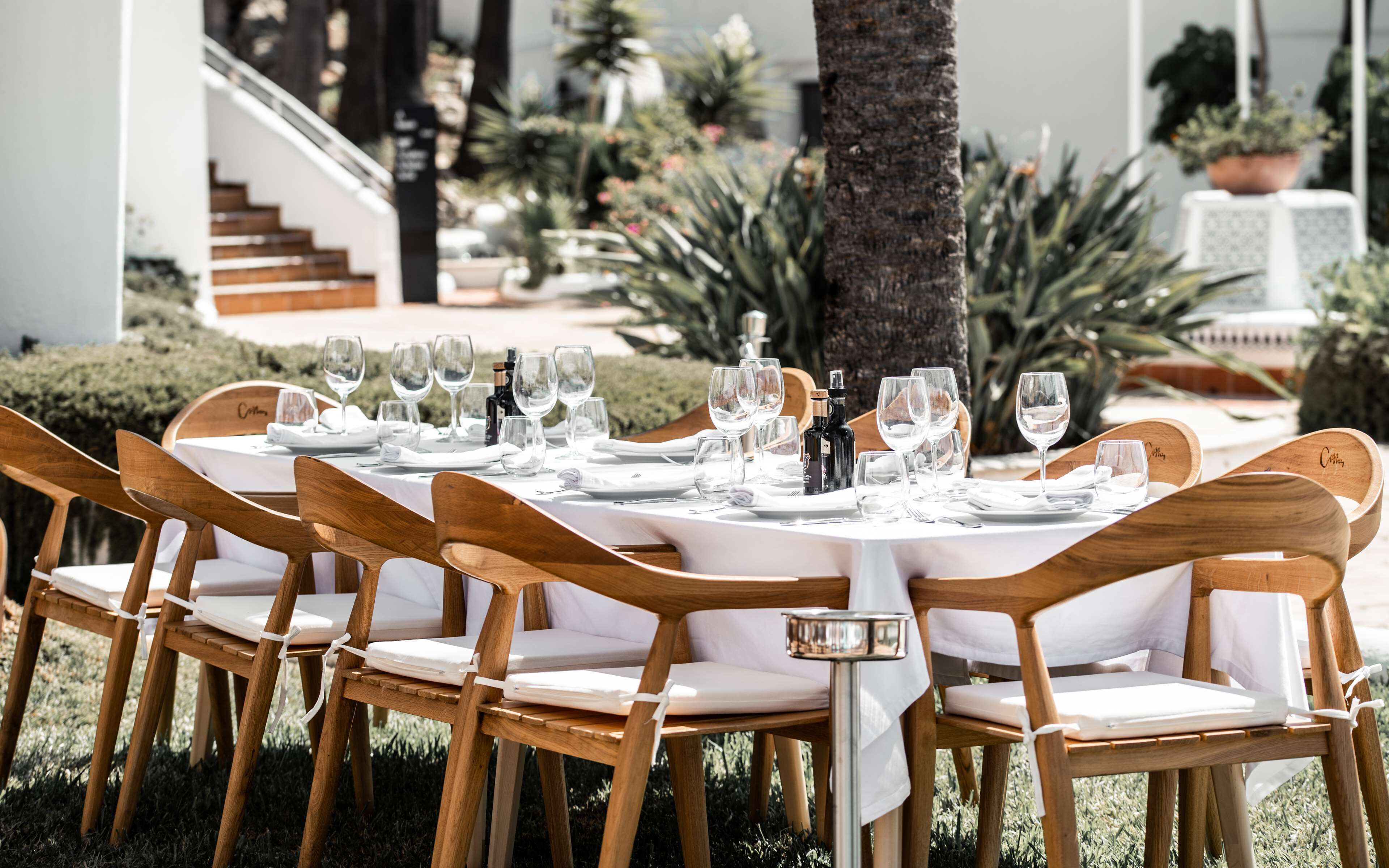 Mesa lista para una comida al aire libre con sillas de madera y manteles blancos.