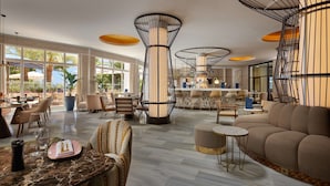 A spacious hotel lobby with a bar area, seating arrangements, and large windows offering views of the outdoors. - Hilton Mallorca Galatzo (Calvia)