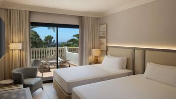 A hotel room with two beds, a chair, a small table, and a balcony with a view of palm trees.