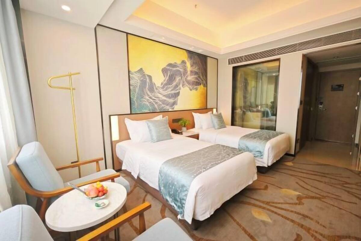 A hotel room with two beds, a small table with fruit, a chair, and a painting on the wall.