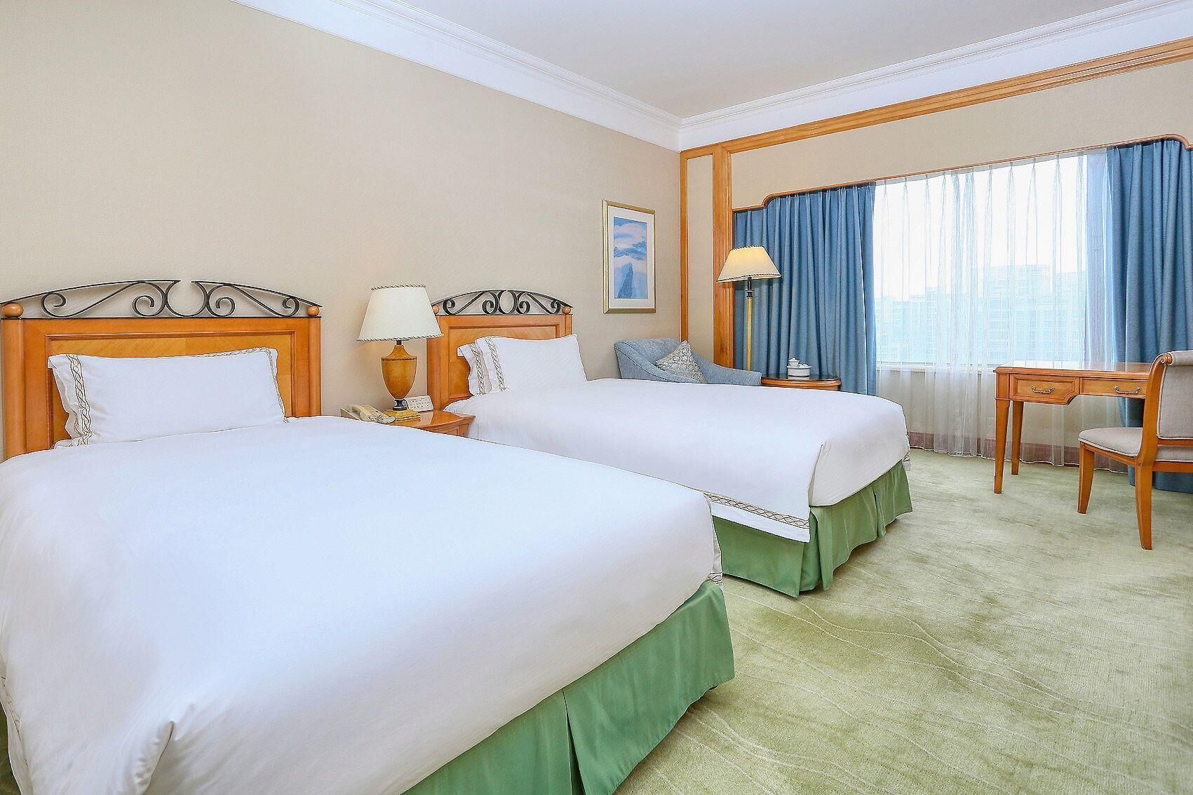 A hotel room with two beds, a desk, a chair, a lamp, and a window with curtains.