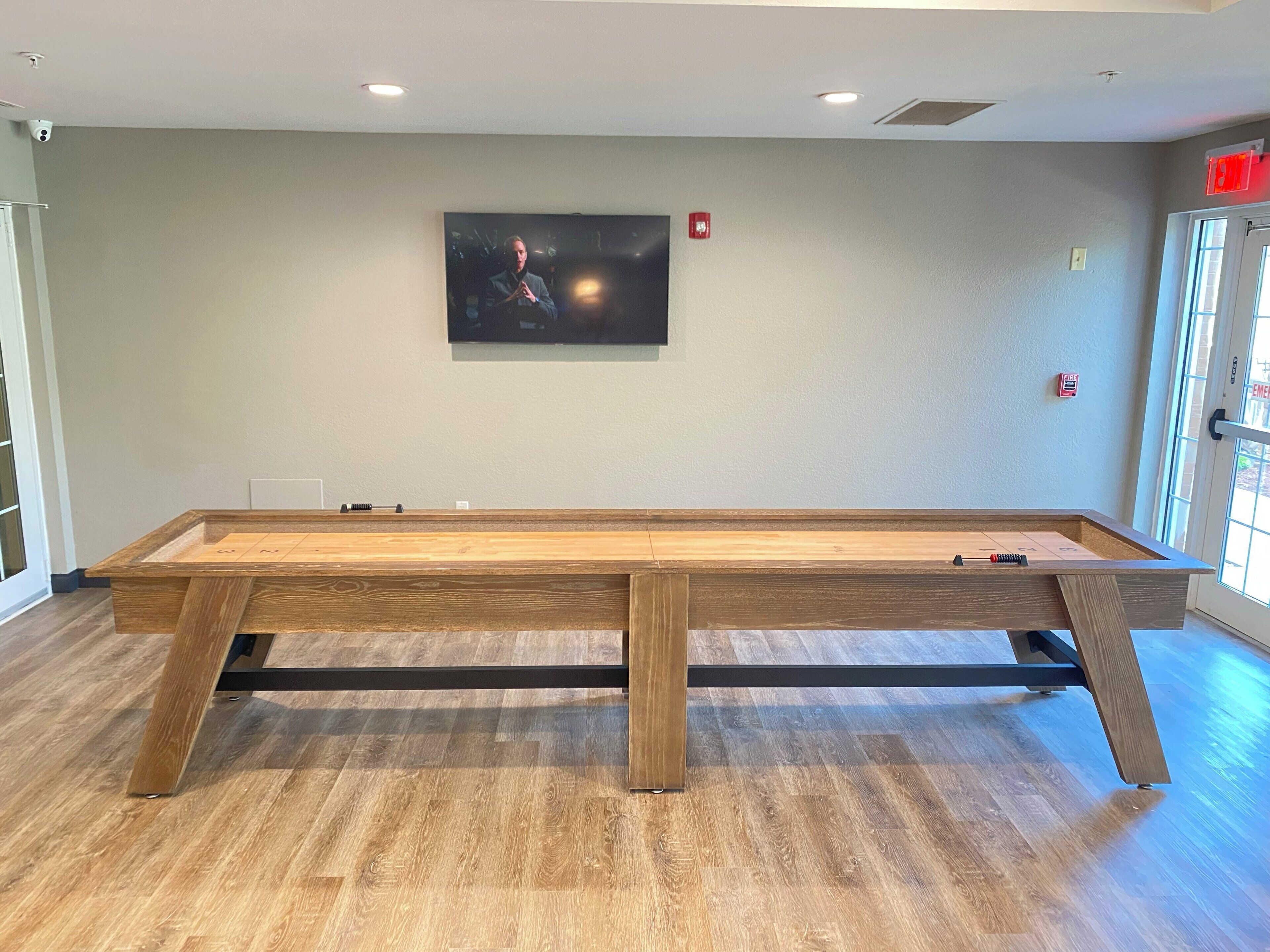 A pool table in a room with wooden flooring, a mounted television on the wall, and a door with an emergency exit sign.