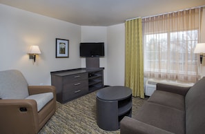 Habitación de hotel con sofá, una mesita, televisor en un mueble bajo, cómoda y ventana con cortinas.