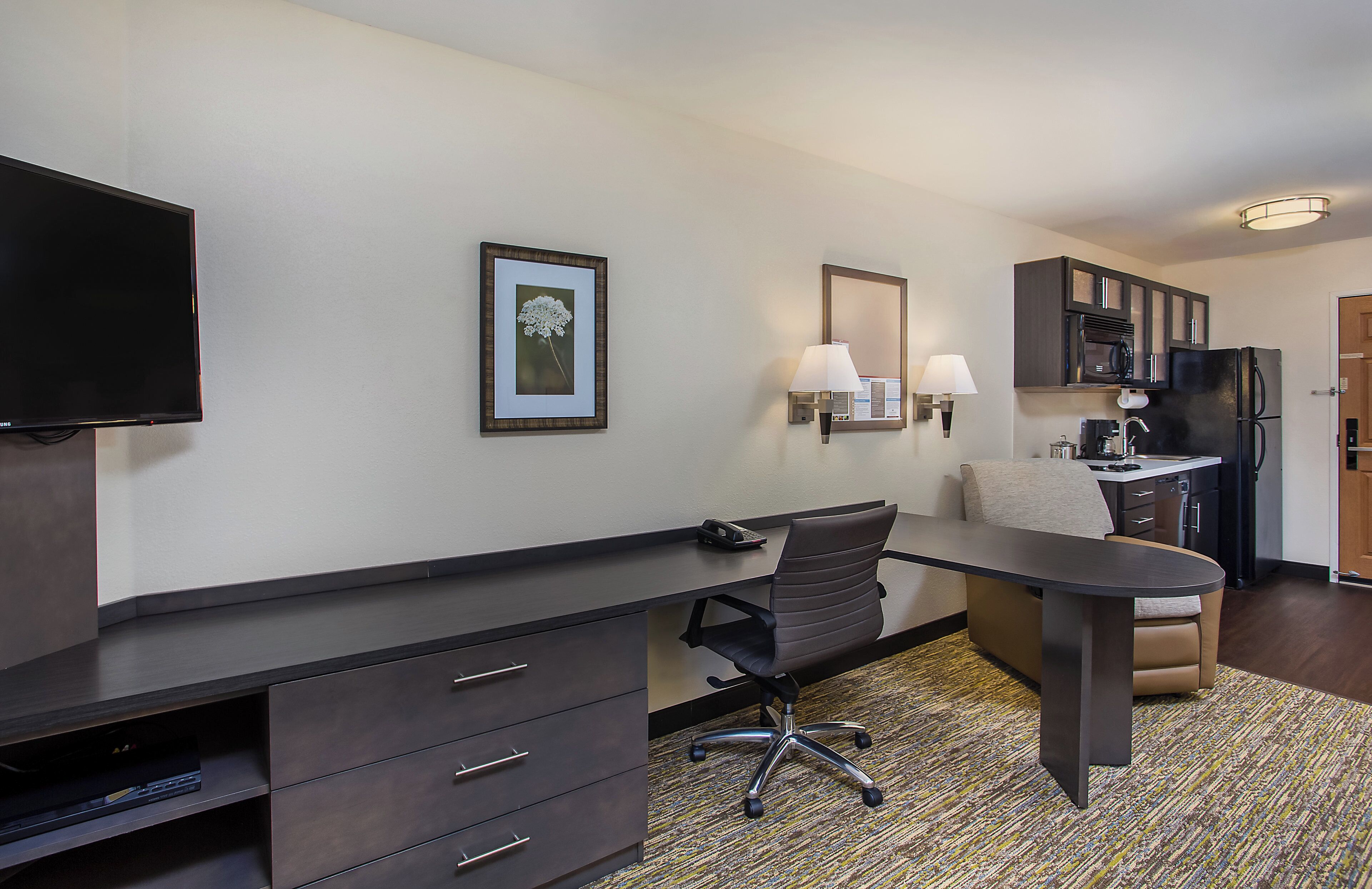 A hotel room with a desk, chair, television, and kitchenette.