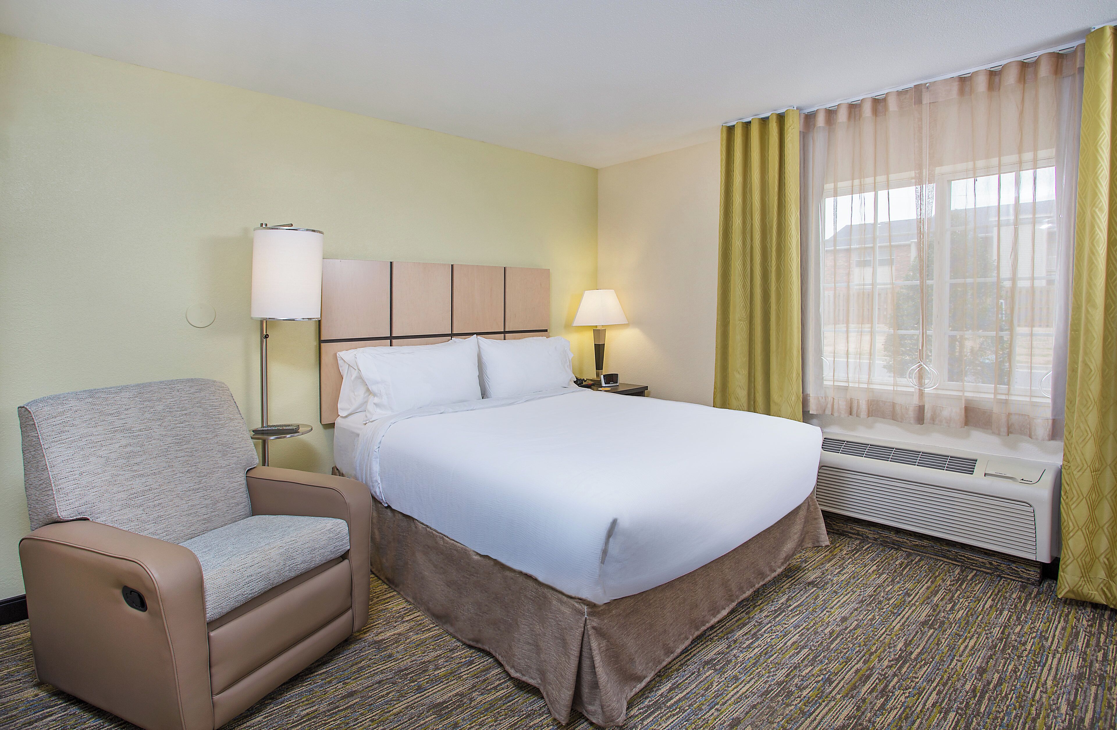 A hotel room with a bed, a chair, a nightstand, a lamp, and a window with curtains.