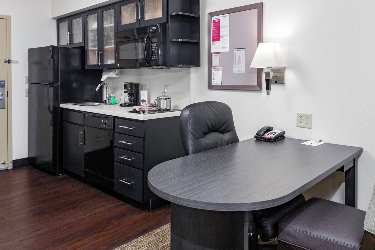 A compact kitchen with a refrigerator, microwave, and sink, a dining table with a chair, and a bulletin board with papers.