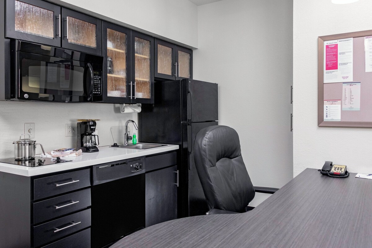 A compact kitchen with a microwave, coffee maker, and refrigerator. There is a dining table with a chair and a phone on it.